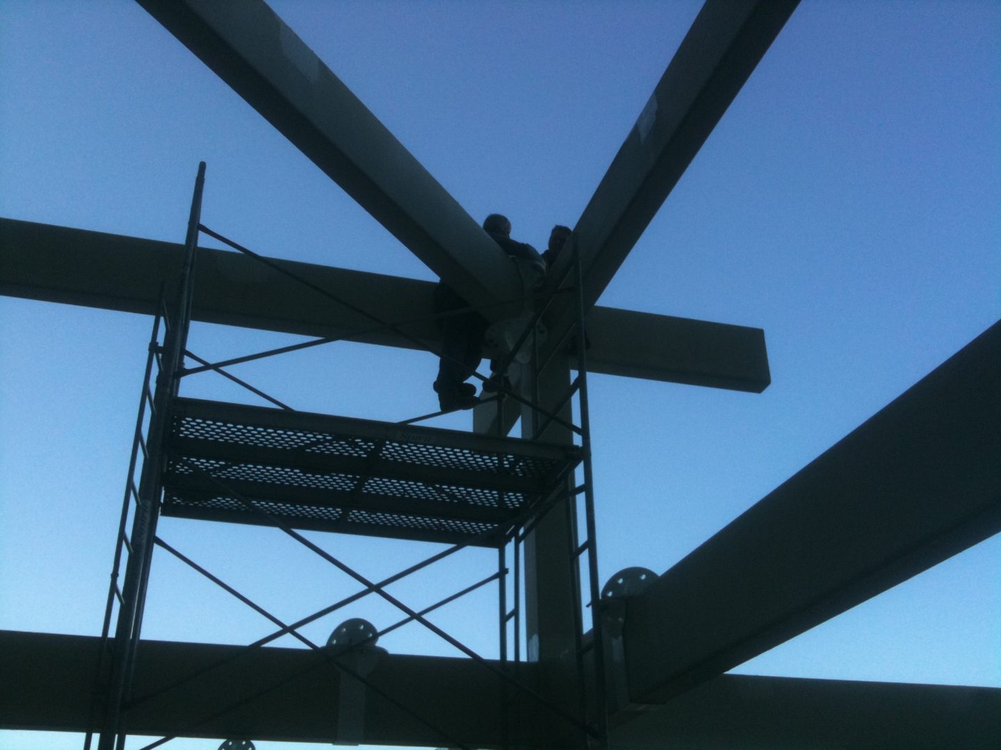 Silhouette of a person working on metal scaffolding against a clear blue sky.