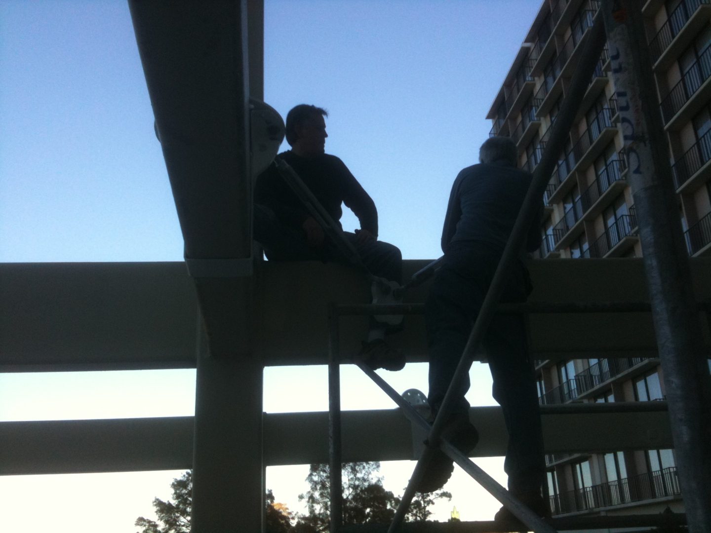 Silhouettes of two people sitting on a beam against a blue sky.